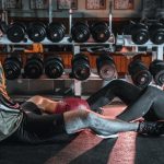A man and woman resting together on the gym floor, surrounded by dumbbells.